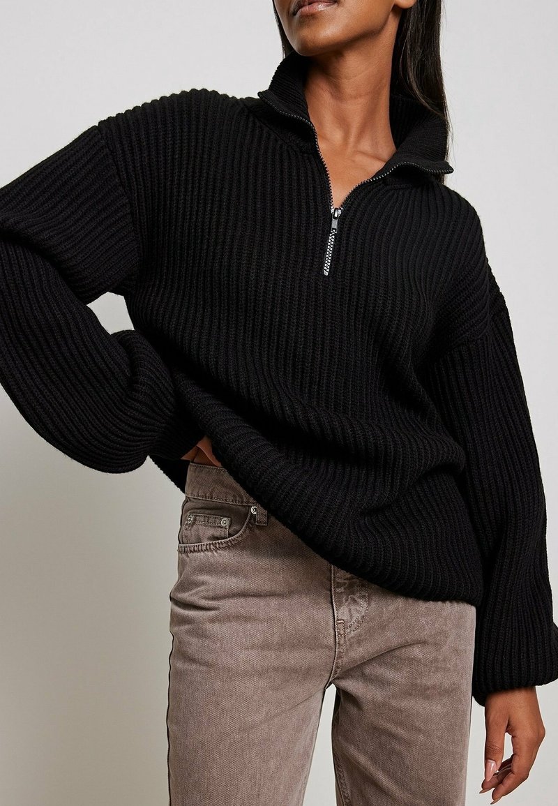 Schwarzer gerippter Pullover mit hohem Kragen und halber Reißverschluss, mit weitem Ärmel. Trägt helle, braune Strukturhosen.