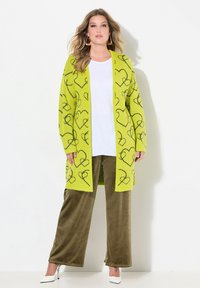 Cardigan verde con motivi a forma di cuore neri, indossato sopra una t-shirt bianca e pantaloni ampi marroni. Caratterizzato da maniche lunghe e un design aperto sul davanti.