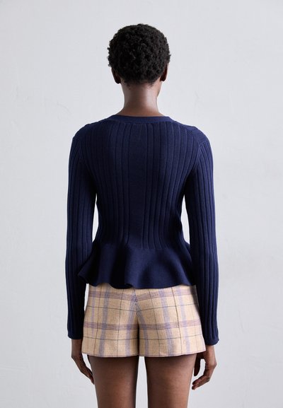 Granatowy sweter z prążkowanym wzorem, z peplum, długimi rękawami i okrągłym dekoltem, połączony z beżowymi szortami w kratę z fioletowymi akcentami.