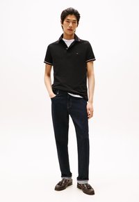 Jovem de pé com as mãos nos bolsos, vestindo uma polo preta, jeans escuros e mocassins castanhos de couro com franjas, sobre fundo branco.