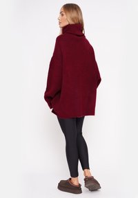 Bordowy, oversize sweter z dzianiny z wysokim kołnierzem, zestawiony z czarnymi legginsami i brązowymi butami wsuwanymi, zaprezentowany od tyłu.