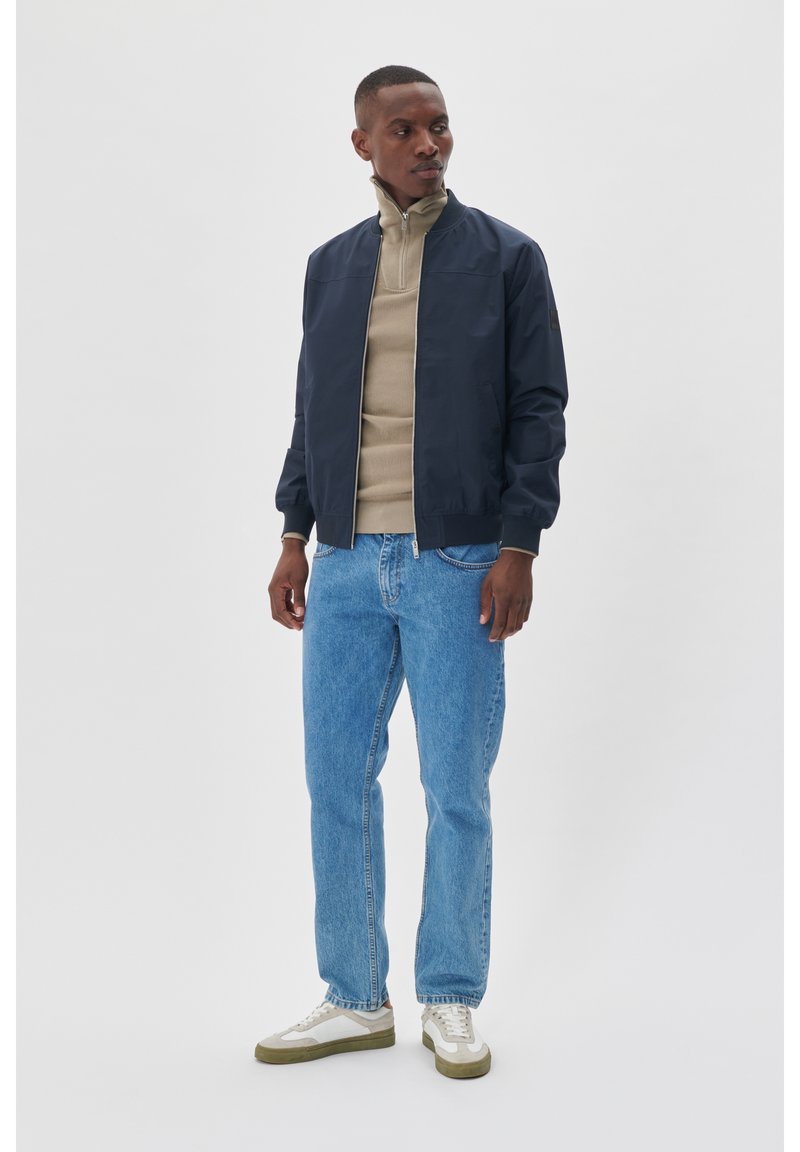 Marine bomberjakke, beige lynlås sweater, lyseblå jeans og hvide sneakers med grønne detaljer. Glatte materialer med en afslappet pasform.