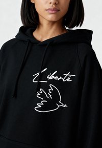 Sweat à capuche noir arborant un texte blanc brodé "Liberté" et un motif de colombe. Le tissu semble doux avec une coupe décontractée.