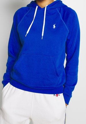 Person trägt einen königsblauen Kapuzenpullover mit weißen Kordeln und kleinem weißen Logo, kombiniert mit weißen Hosen mit rotem Logo nahe der Tasche.