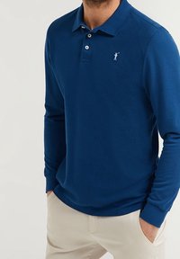 Långärmad polo-shirt i marinblått, med texturerad tyg, treknappsknäppning och en vit logotyp på vänster bröst.