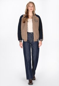 Fleecejakke i tan og marineblå, med ribbede mansjetter og krave, lynlås lukning og broderet tekst. Kombineret med højtaljede blå jeans.