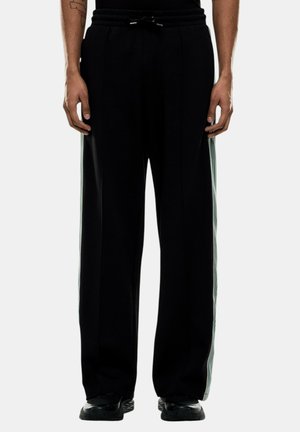 Uomo che indossa pantaloni neri a gamba larga con strisce laterali bianche e sneakers nere, in piedi contro uno sfondo chiaro uniforme.