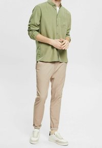 Camicia verde chiaro con bottoni e tasca frontale, abbinata a pantaloni beige a vita alta e sneakers bianche con dettagli grigi.