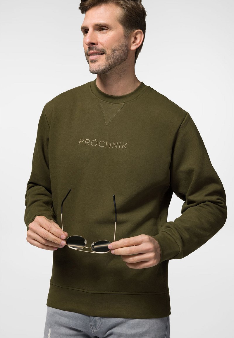 Oliwkowa bluza z okrągłym dekoltem, teksturowana tkanina i wyszywane logo. Noszący trzyma czarne okulary przeciwsłoneczne w złotej oprawie.