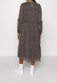 Kobieta ubrana w czarną sukienkę midi z różowym kwiatowym nadrukiem, z długimi przezroczystymi rękawami i białymi trampkami w skarpetkach, stojąca na białym tle.