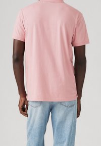 Polo à manches courtes, rose uni, avec un ourlet droit et des fentes latérales. Le tissu semble doux avec une coupe décontractée, associé à un jean bleu clair.