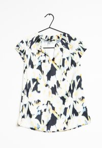 Blouse à manches courtes avec un motif abstrait noir, blanc, jaune et bleu, accrochée à un cintre noir sur un fond blanc.