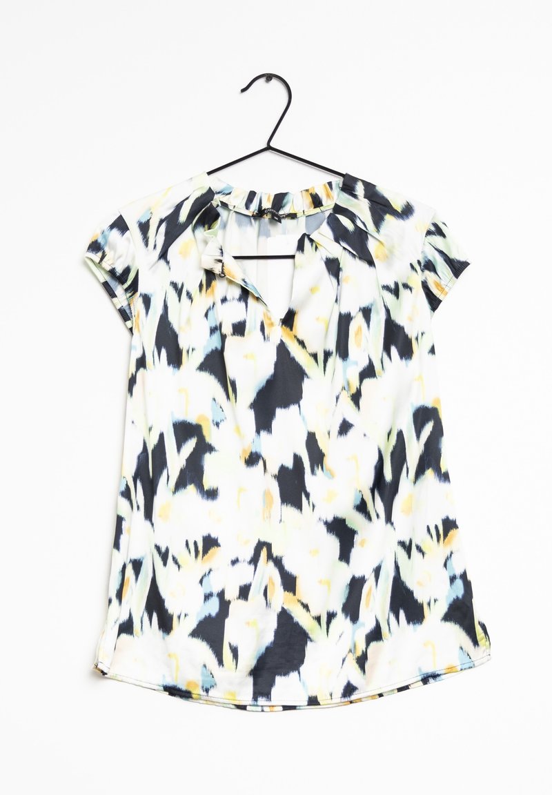 Blouse à manches courtes avec un motif abstrait noir, blanc, jaune et bleu, accrochée à un cintre noir sur un fond blanc.