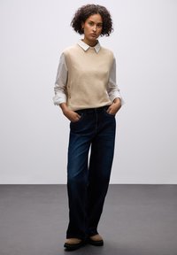 Beige gebreide vest over een wit overhemd met kraag, gecombineerd met donkerblauwe wijde jeans en bruine suède enkellaarsjes. Eenvoudig, schoon ontwerp.