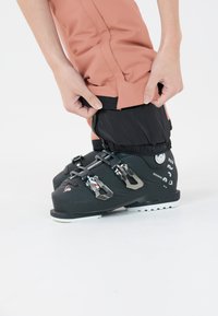 Zwarte skibotten met metalen gesp, gedragen met roze waterdichte broek. De broek heeft een zwart elastisch manchet dat wordt aangepast.