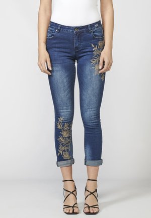 Frau trägt dunkelblaue, hochgekrempelte Skinny-Jeans mit goldener Blumenstickerei am Oberschenkel und Wadenbereich, kombiniert mit schwarzen Riemchenabsätzen.