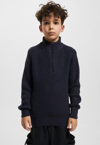 Jūras spēku adīts džemperis ar augstu apkakli un rāvējslēdzēju, ar faktūru rakstu un ribotām piedurknēm, uzvilkts uz melnajām biksēm.