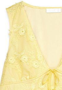 Yellow sleeveless dress with floral embroidery, deep V-neckline, and a decorative tie at the front. Soft, sheer fabric with delicate details.