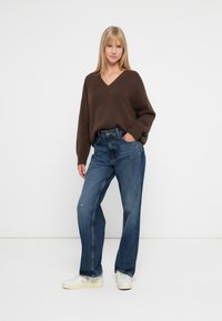 Brun oversized v-hals genser, blå høytlivsjeans og hvite joggesko. Enkelt design, myk tekstur og avslappet passform.