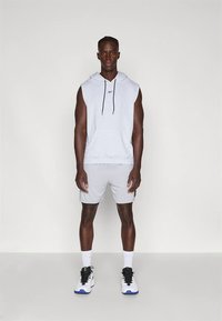 Άνδρας φορεί ανοιχτό γκρι αμάνικο φούτερ, ταιριαστά σορτς, λευκές κάλτσες και λευκά αθλητικά παπούτσια με μαύρη κορδόνια, και στέκεται μπροστά από απλό φόντο.