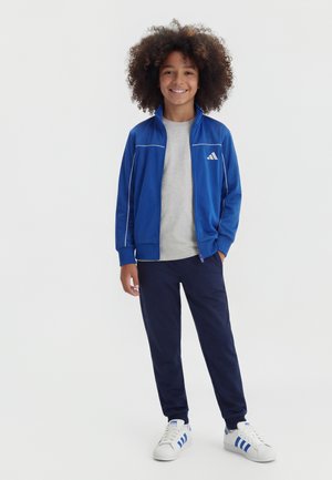 Smilende barn med krøllet hår iført blå lynlåstrøje, grå skjorte, marineblå bukser og hvide sneakers med blå striber, stående med hænderne i lommerne.