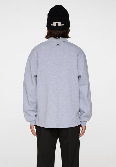 Osoba ubrana w jasnoszarą koszulę z długim rękawem i czarne spodnie, stojąca tyłem, z czarną dzianą czapką z białym wzorem.