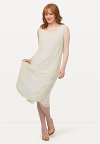 Vestido sin mangas color crema con un sutil bordado floral amarillo, diseño en forma de A y cuello redondo, combinado con sandalias doradas con tiras.
