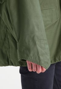 Jachetă utilitară verde cu o textură netedă, cu manșete pliate și o buzunar vizibil, însoțită de blugi închiși la culoare.