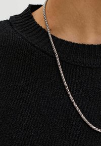 Plano detalle de una persona que lleva un collar de cadena plateada sobre un suéter negro de punto con cuello redondo.