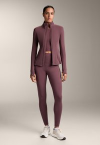 Lila idrottsjacka med hög krage, kombinerad med en matchande kort topp och leggings. Texturerat tyg och figursydd design. Vita sneakers.