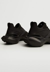 Chaussures de sport noires présentant une tige en mesh texturé, une semelle épaisse et rainurée, et un design minimaliste. L'arrière affiche un logo discret.