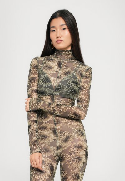 Mujer con cabello largo y liso que lleva un bodysuit floral de alto cuello y transparente con grandes flores beige y verdes, posando con un brazo cruzado sobre su cuerpo.