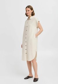 Abito button-up, lunghezza al ginocchio, in tessuto bianco sporco, con maniche corte, tasche laterali e orlo curvo; presenta bottoni marroni scuri. Indossati mules nere.