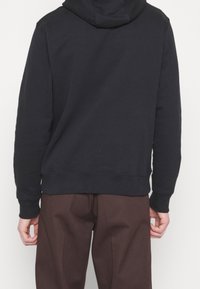 Marinblå huvtröja med avslappnad passform, ribbade muddar och fåll; bärs med bruna byxor. Mjuk textur, minimalistisk design utan synliga logotyper.