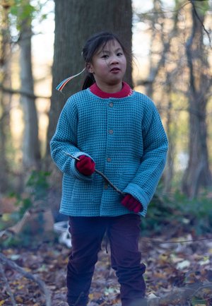 Blaue strukturierte Strickjacke, roter Kragen, rote Handschuhe, dunkle Hose, hält einen Stock, in einer Waldumgebung mit Laub auf dem Boden.