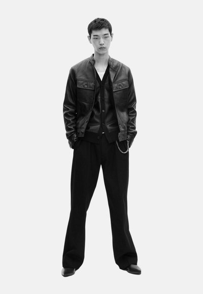 Chaqueta de cuero negra con dos bolsillos frontales, sobre un cárdigan negro. Pantalones sueltos negros y zapatos negros completan el conjunto.