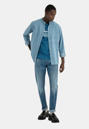 Camisa de jeans azul claro con cuello redondo, camiseta verde azulado debajo con rayas blancas y texto, jeans azul claro y zapatos negros con suela gruesa.