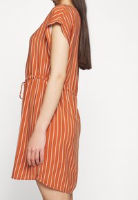 Rostfärgad klänning med vita vertikala ränder, korta ärmar, dragsko i midjan och en lös passform. Nedtill är längden något rundad.