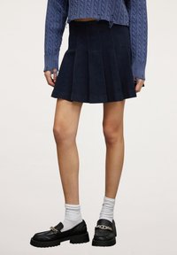 Námornická skládací sukně vyrobená z měkké látky, kombinovaná s černými mokasíny s řetízkovým akcentem a bílými ponožkami, pod vzorovaným modrým svetrem.