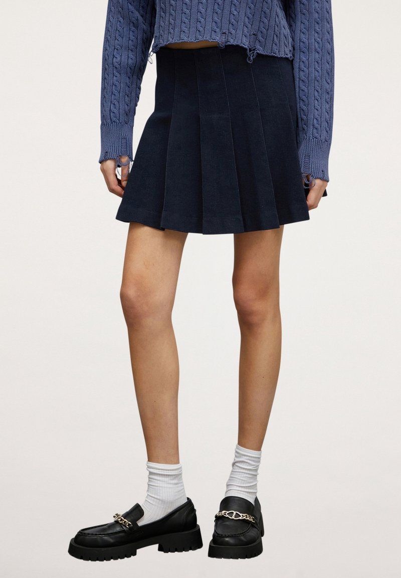 Námornická skládací sukně vyrobená z měkké látky, kombinovaná s černými mokasíny s řetízkovým akcentem a bílými ponožkami, pod vzorovaným modrým svetrem.