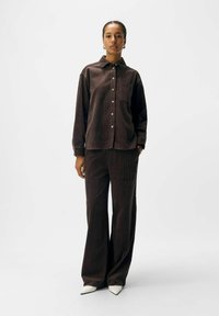 Woman stands wearing dark brown corduroy button-up shirt and matching wide-leg pants with white pointed shoes against a plain background.