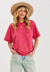 Różowy sweatshirt z krótkimi, podwiniętymi rękawami, zaokrąglonym dołem i logotypem na piersi. Połączony z jasnoniebieskimi spodniami o wysokim stanie. Beżowy kapelusz z szerokim rondem.