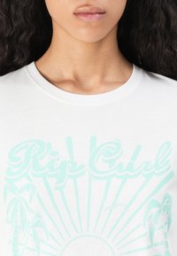 Detailný záber na osobu v bielej košeli s svetlomodrým textom "Rip Curl" a grafikou palmy a slnka na hrudi.