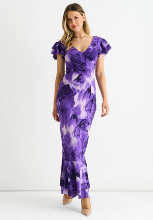 Woman wearing a fitted purple floral maxi dress with ruffled short sleeves and beige high heels, standing against a plain white background.