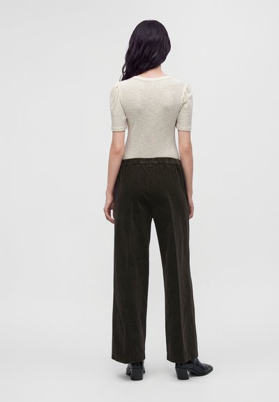 Femme aux longs cheveux foncés portant un haut beige à manches courtes et un pantalon noir ample, debout, dos tourné, sur un fond clair uni.