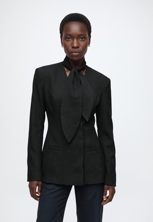 Femme aux cheveux courts porte un blazer noir cintré avec une écharpe oversize nouée au cou et un pantalon sombre sur fond blanc uni.