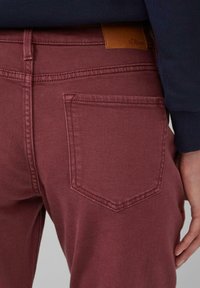 Burgundi denimpüksid, millel on voolav lõige, tagakotis ja pruuni naha brändipatchi küljes. Sile tekstuur.
