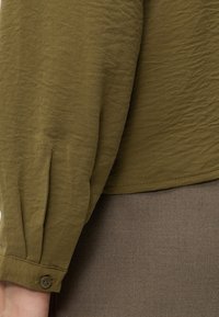 Gros plan d'une personne portant une chemise à manches longues vert olive avec des poignets boutonnés et un pantalon marron texturé.