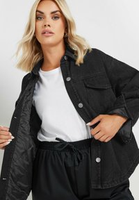 Musta denimhame, jossa on napsautettavat napit ja etutaskut, kerroksittain valkoisen t-paidan päällä. Kuoletettu kangas, vuoraamaton sisus, rento istuvuus.