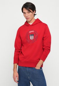 Sweatshirt vermelho com um gráfico redondo de um desenho de escudo nas cores azul marinho, branco e vermelho. Confeccionado em material suave, possui um bolso canguru.